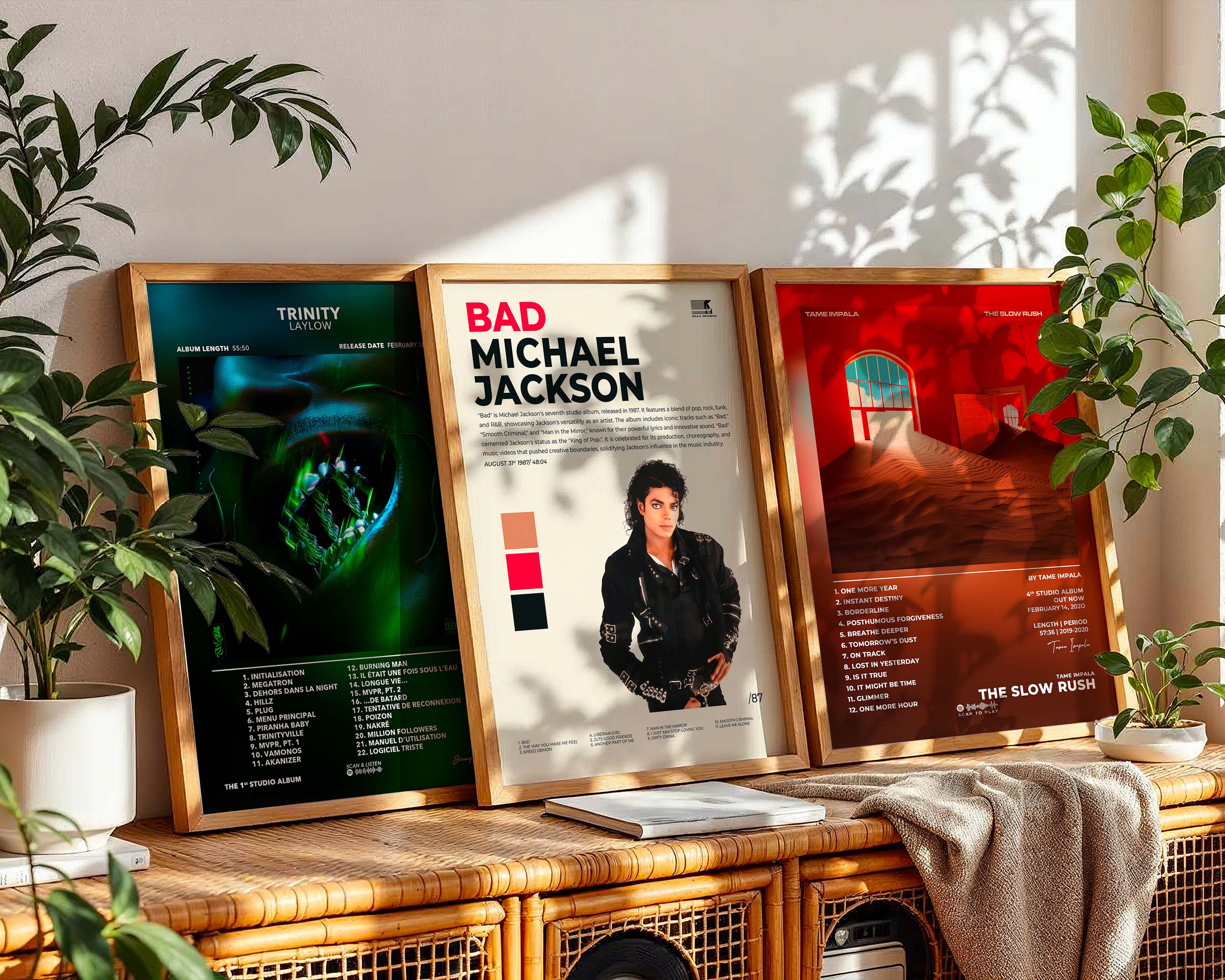 Trois posters d'albums musicaux encadrés en bois (Trinity de Laylow, Bad de Michael Jackson et The Slow Rush de Tame Impala) alignés sur un meuble en rotin à côté de plantes vertes.