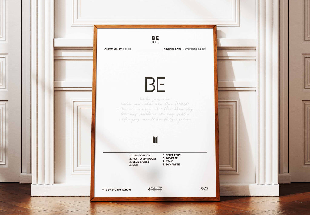 Affiche encadrée de l'album BE de BTS posée au sol contre un mur pour une décoration intérieure douce et moderne.