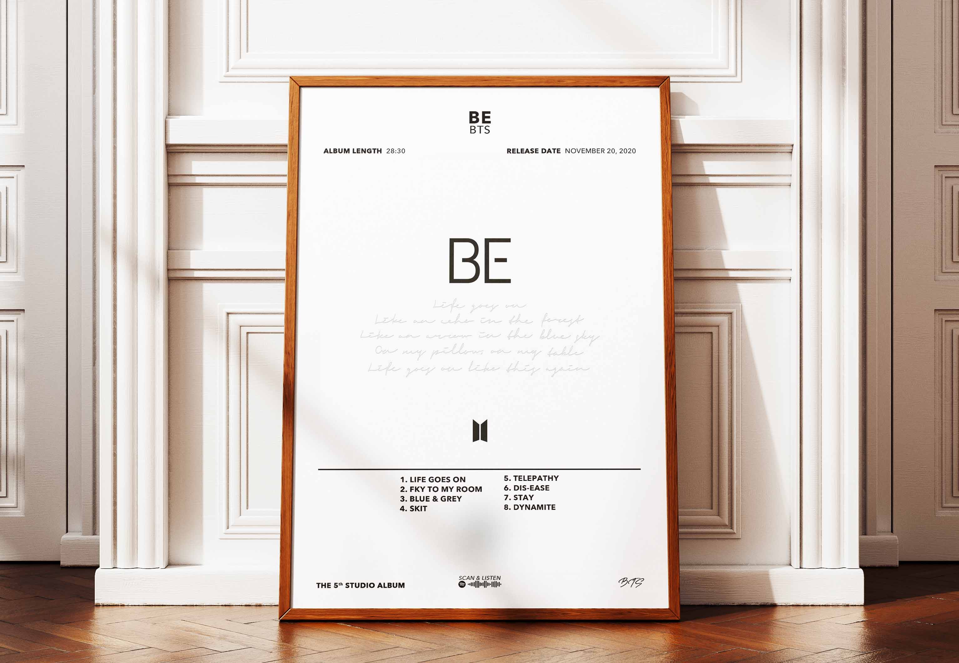 Affiche encadrée de l'album BE de BTS posée au sol contre un mur pour une décoration intérieure douce et moderne.
