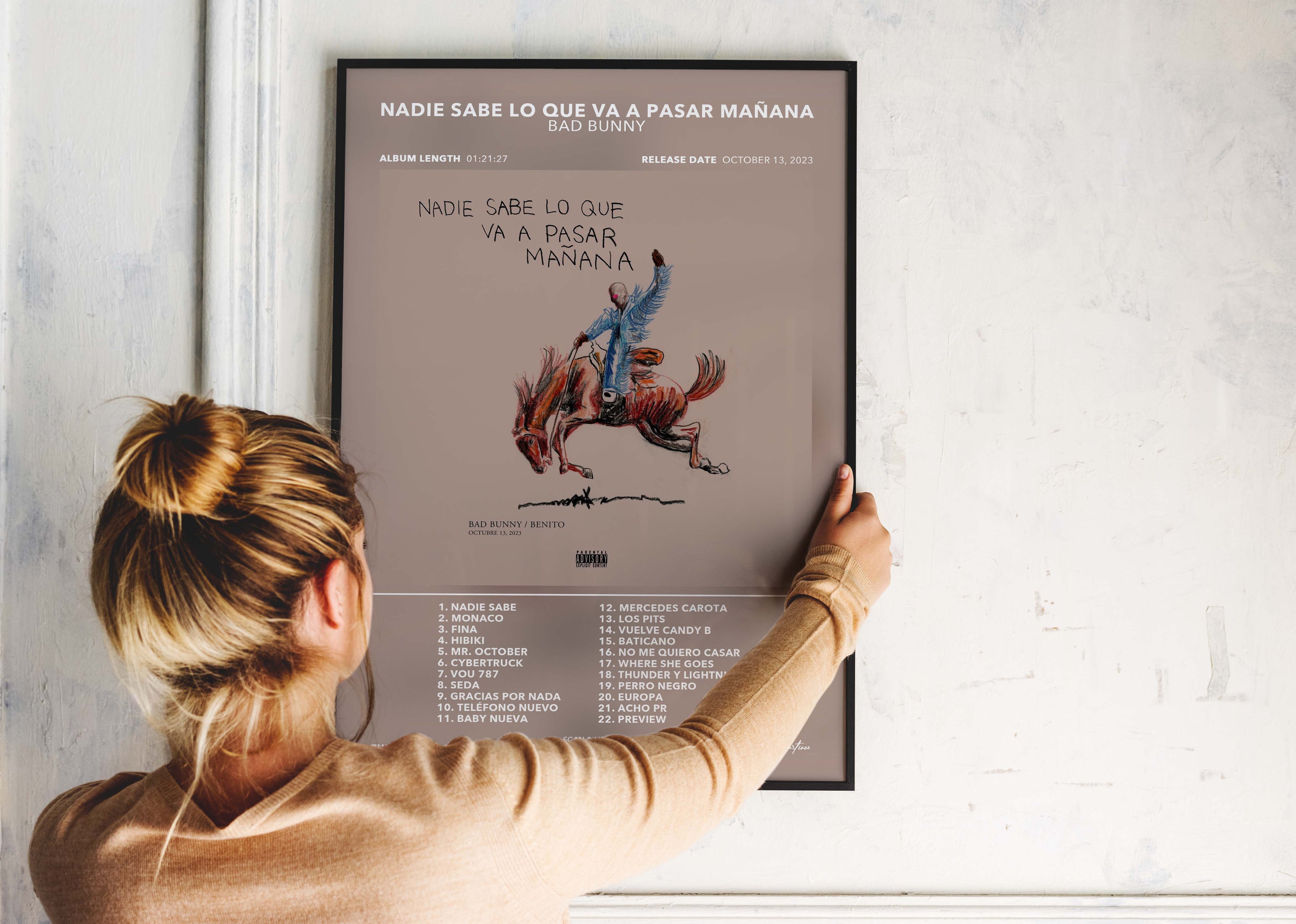 Une personne installe le poster encadré de Bad Bunny Nadie Sabe Lo Que Va A Pasar Mañana sur un mur blanc de salon.