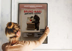 Une personne installe le poster encadré de Bruno Mars Unorthodox Jukebox sur un mur blanc pour personnaliser son espace.