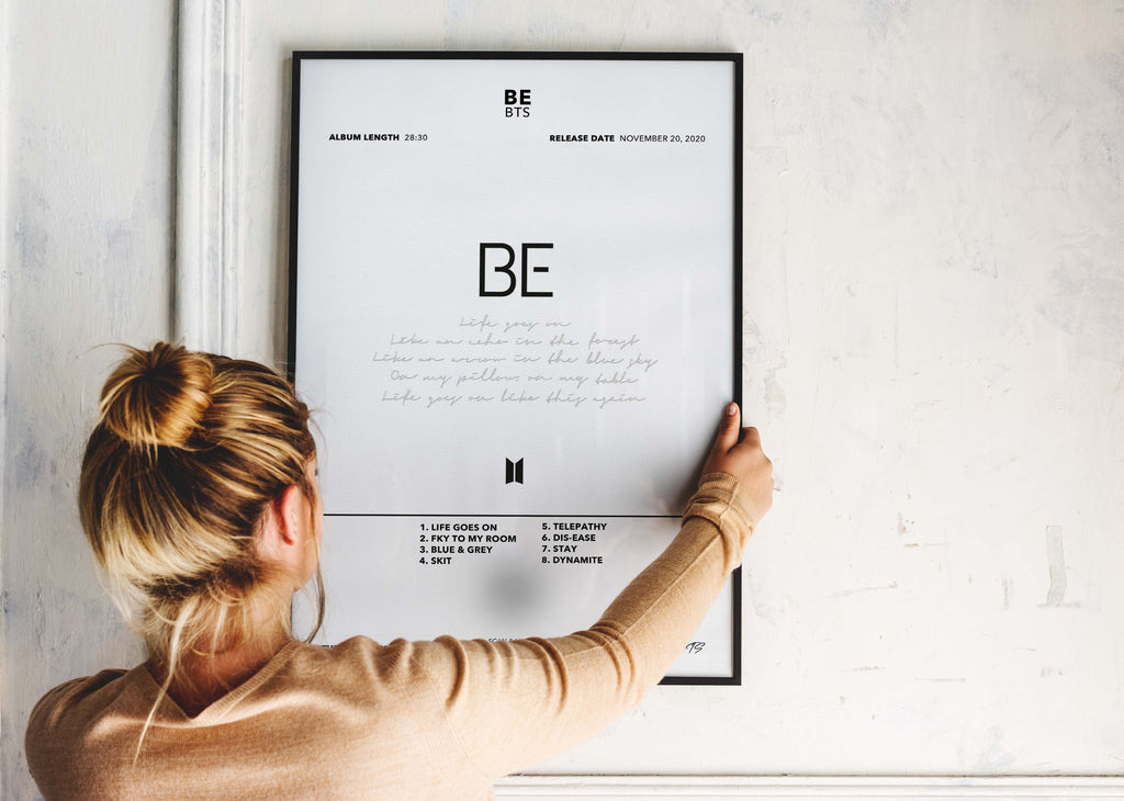 Une personne installe le poster encadré de BTS BE sur un mur blanc pour personnaliser son espace avec élégance.
