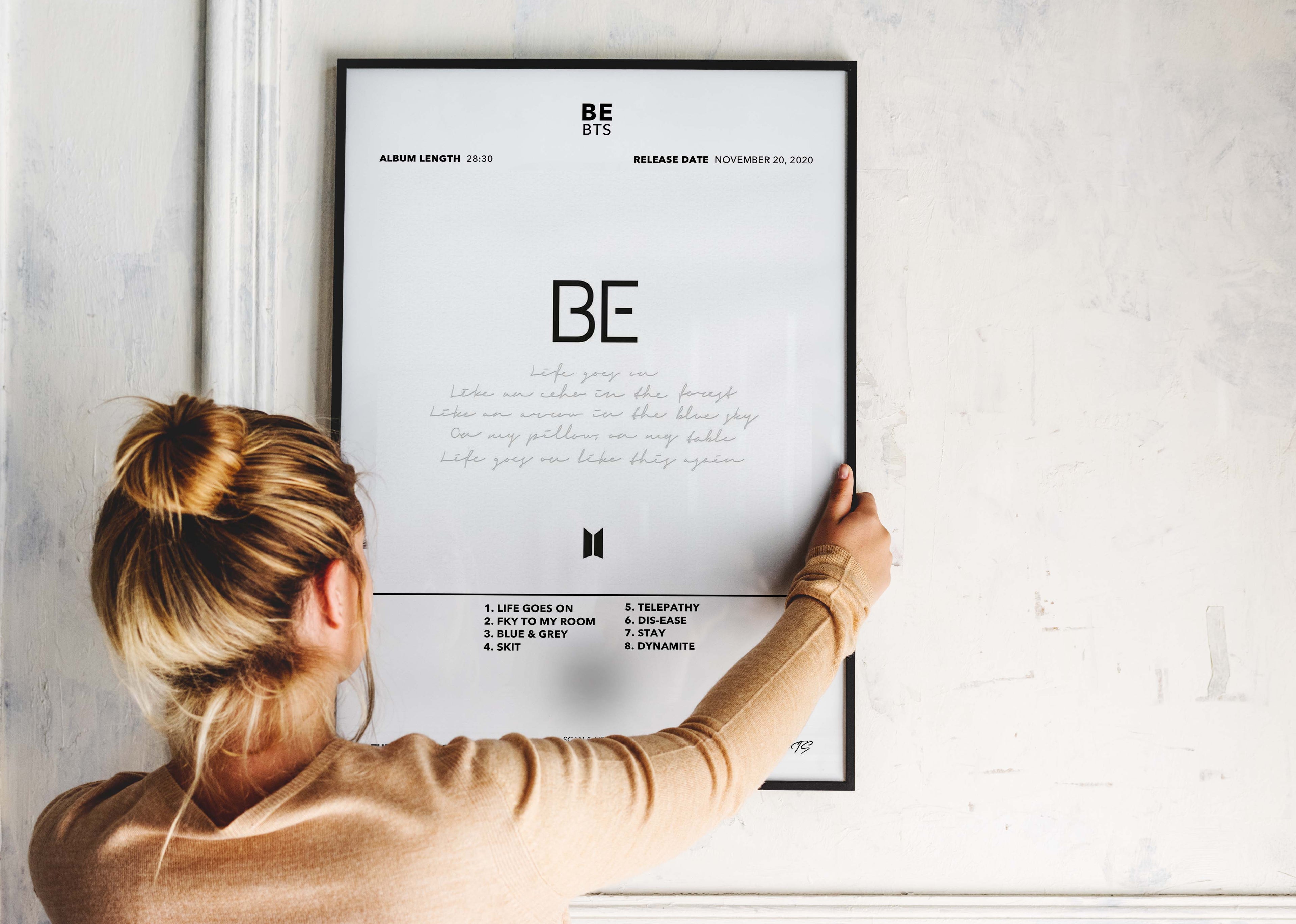 Une personne installe le poster encadré de BTS BE sur un mur blanc pour personnaliser son espace avec élégance.
