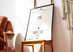 Cadre contenant l'affiche de l'album BE de BTS posé sur une chaise design dans un salon lumineux au style épuré.