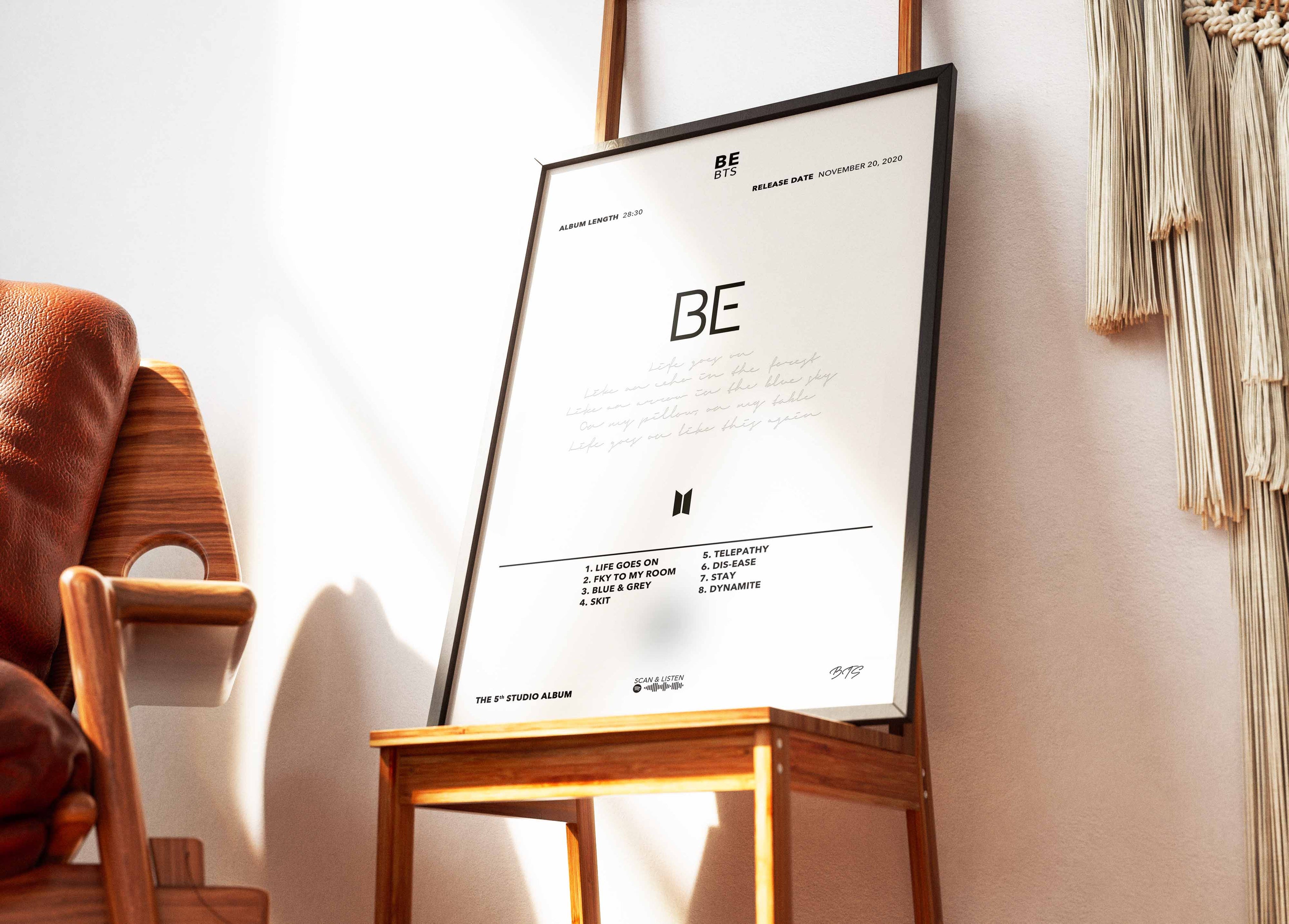 Cadre contenant l'affiche de l'album BE de BTS posé sur une chaise design dans un salon lumineux au style épuré.